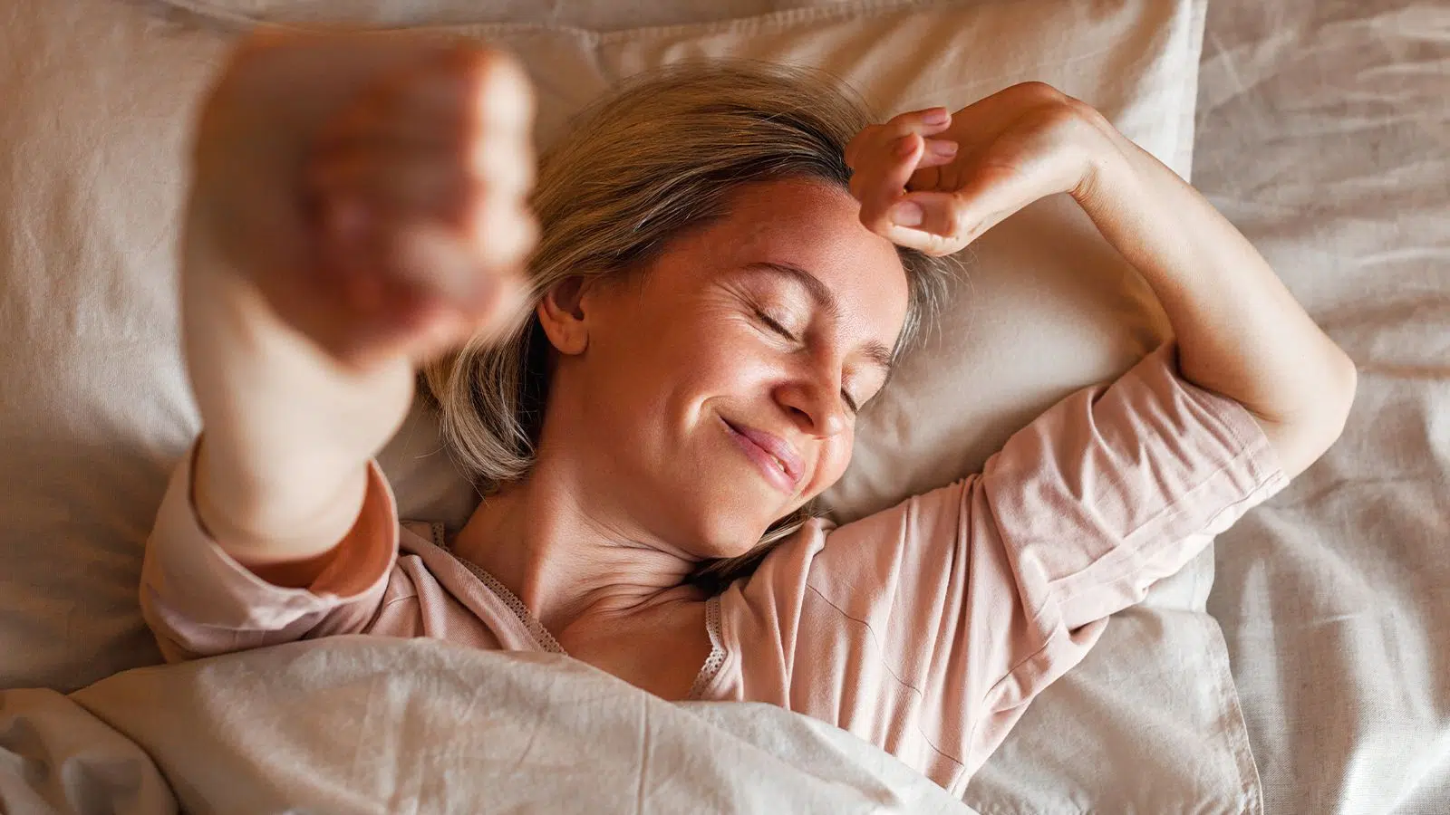 A woman lying in bed, stretching her arms and smiling, looking relaxed.